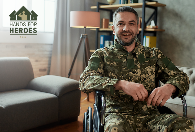 a smiling soldier wearing military fatigues sits in a wheelchair inside his home next to the Hands for Heroes logo